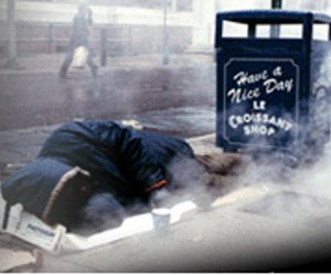 homeless man in blue coat lying next to a black trash can that says "Have a Nice Day Le Croissant Shop"